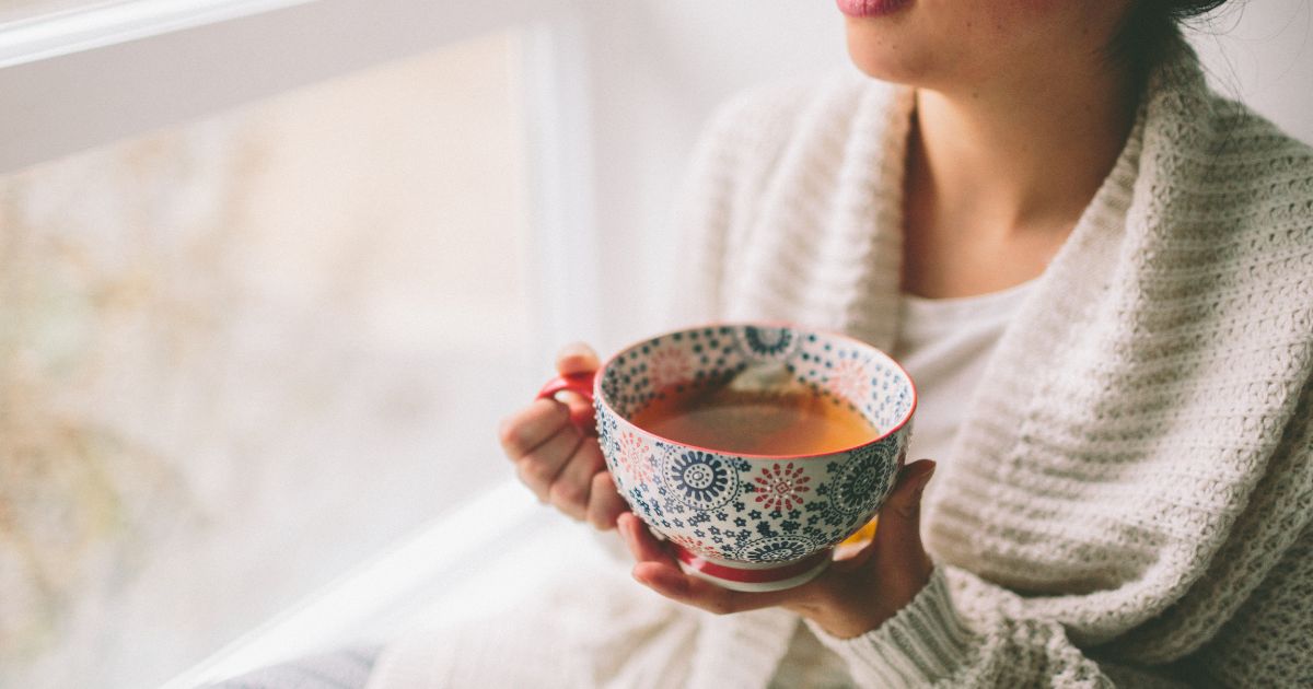 A woman wearing a thick cardigan while holding a cup of tea as she stands beside a window.