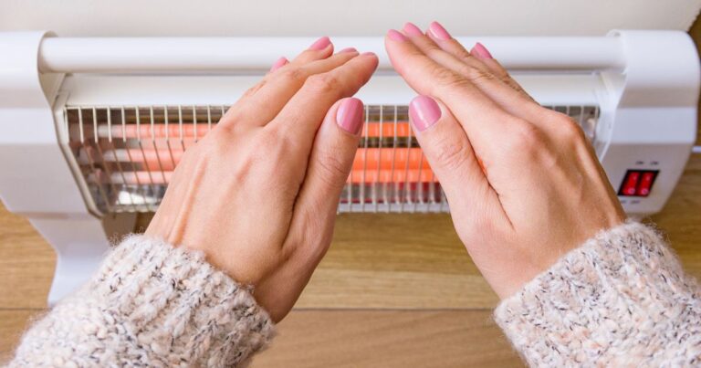Manicured hands wearing a knitted jumper, trying to catch heat from a heater against the wall.