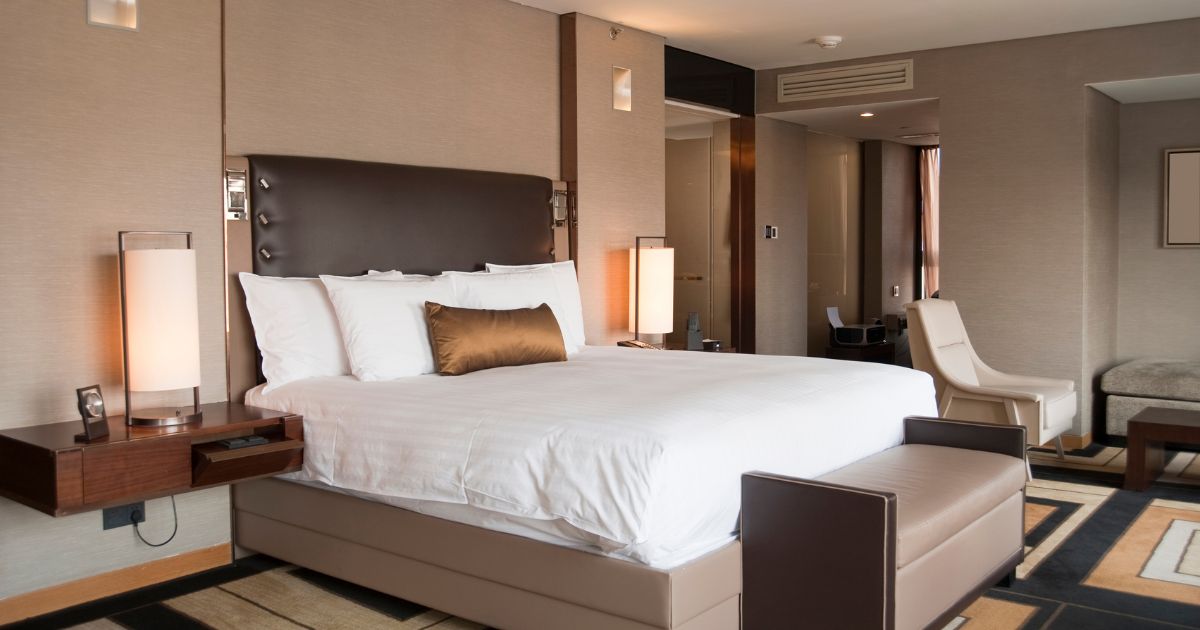 A hotel room with a queen sized bed, two side tables and lamps on both sides, a white single sofa and an ottoman in front of the bed.