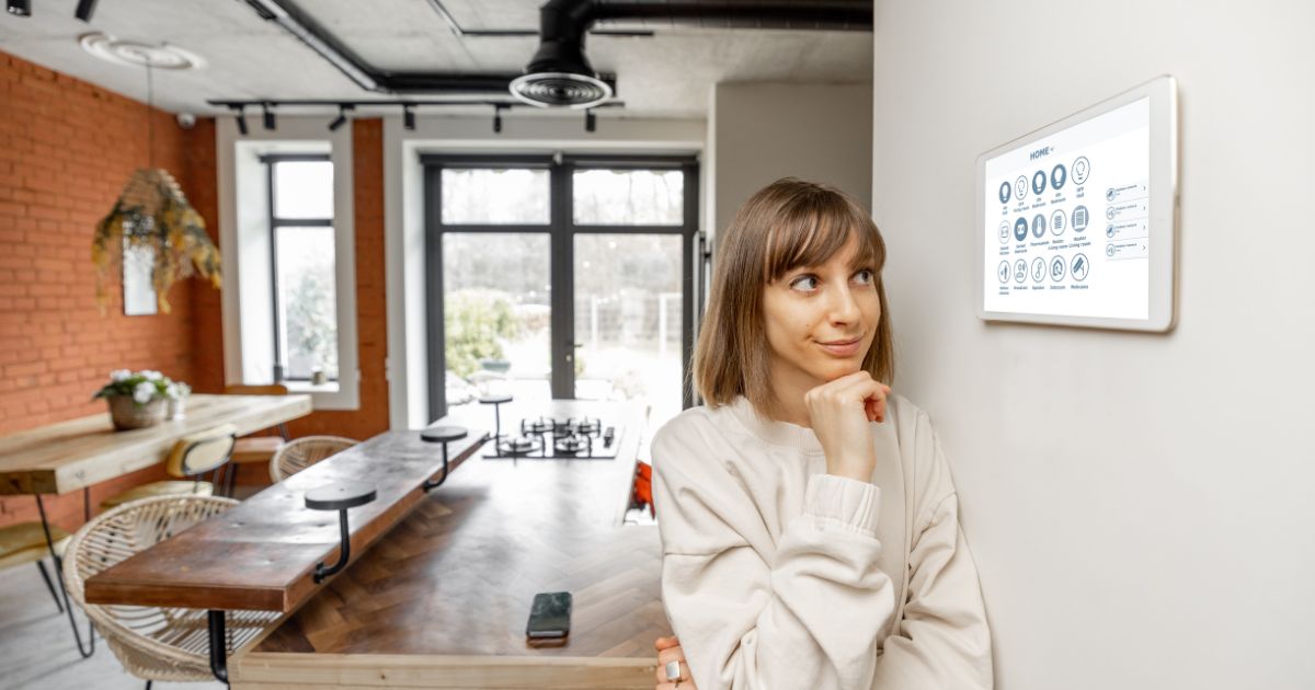 A woman looking at a wall mounted thermostat as if thinking about adjusting the temperatures. Behind her is a modern looking dining area with glass doors at the far end of the room leading outdoors. Outside, the trees are covered with snow.