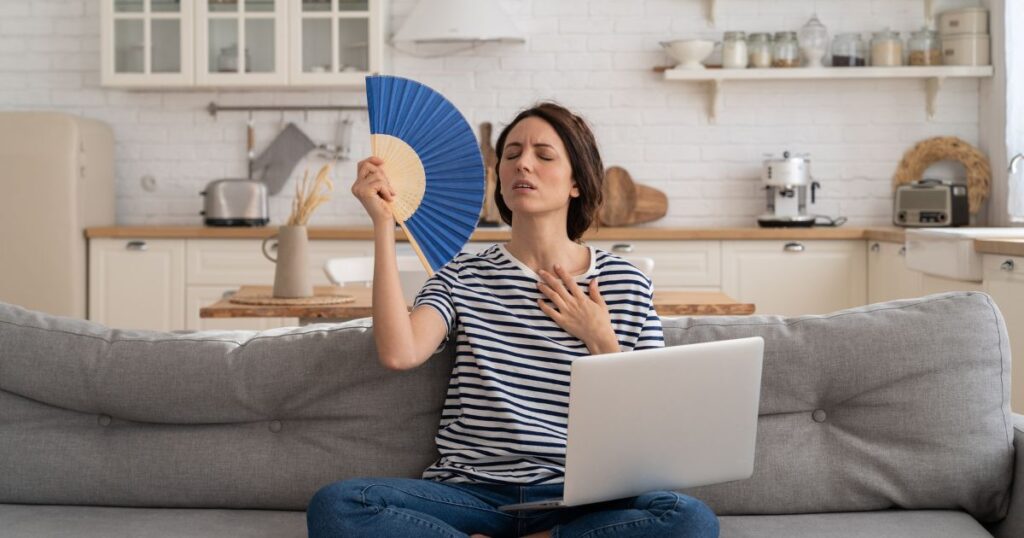 A woman wearing a stiped t-shirt and jeans, fanning herself while a silver laptop sits on her lap. The woman appears to be feeling so much heat and looks distressed as she sits on a gray couch with a view of a kitchen with minimal appliances and decors in it.