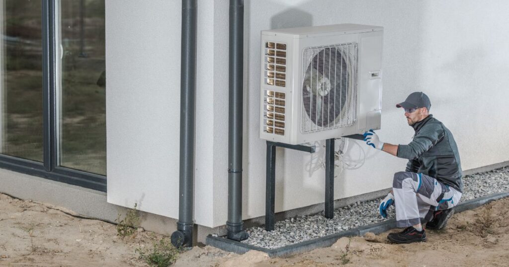 A man performing repairs on a heat pump placed on metal stilts outdoors. 