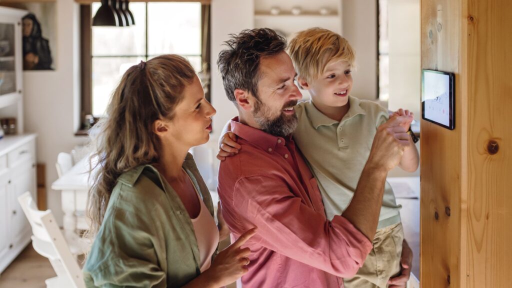 A family of 3 in a kitchen. The father is seen carrying his son as they adjust the thermostat with the mother standing behind the father. Their home is equipped with a heating and cooling system maximising their savings via the Victorian Energy Upgrade (VEU) Program.