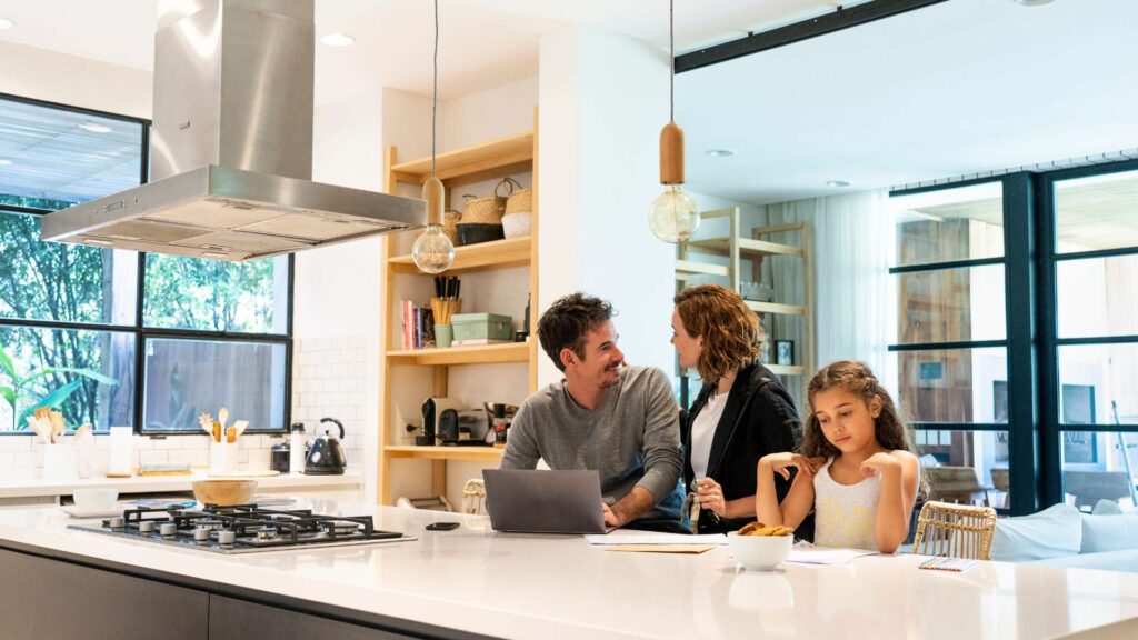 A family of three sitting along a kitchen counter top. The mother and father are happily talking as the father works on his laptop in the kitchen island, whilst their daughter in looking down on the countertop.
