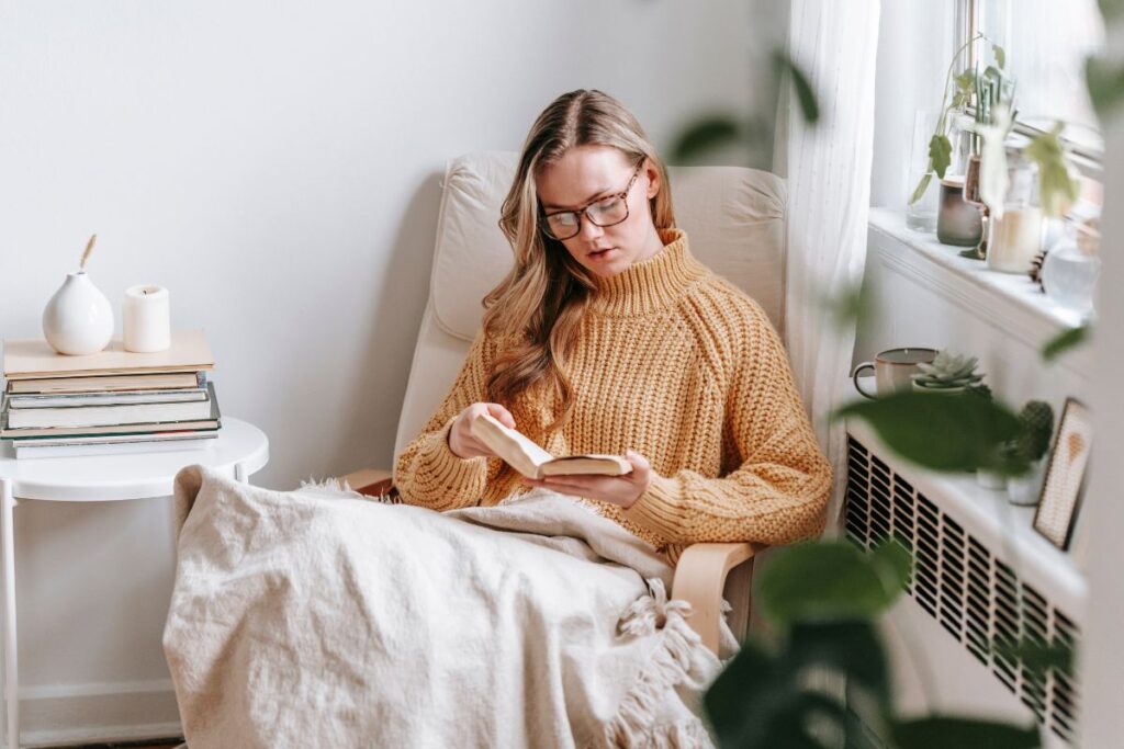 A woman in a beige jumper, sitting cosily in a sofa near a window, with a book and a blanket in her lap. Bundling up and taking advantage of daylight heat is one of the ways to avoid heating mistakes that cost you money in winter.