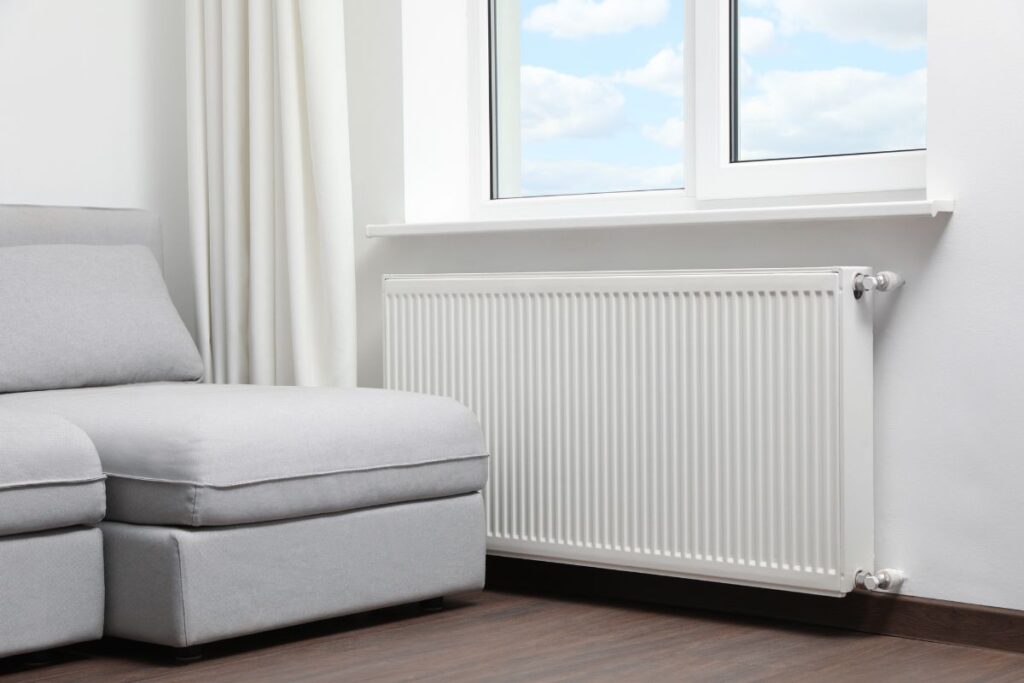 A wall-mounted heater installed in a living room, just beneath a window. Next to the heater in a gray sofa.