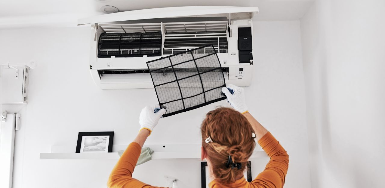 A woman in an orange overall replacing the filter in a split type air conditioner. The value of a professional air conditioner cleaning service lies in the prolonged lifespan of an air conditioning unit.
