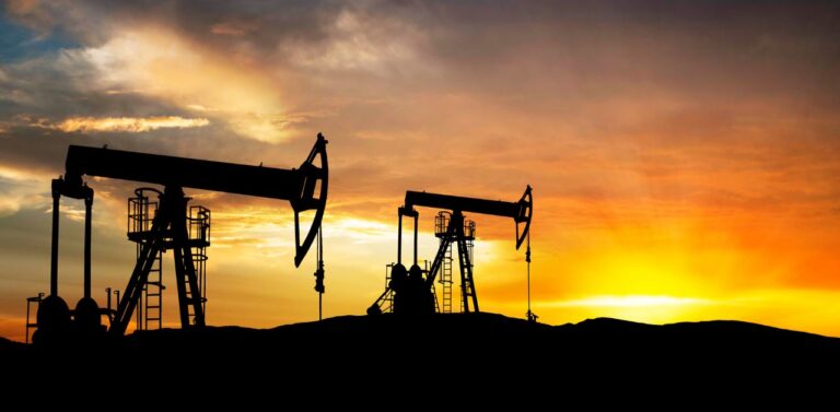 A silhouette of two oil pumpjacks extracting crude oil from the ground. The image is taken at sunset.