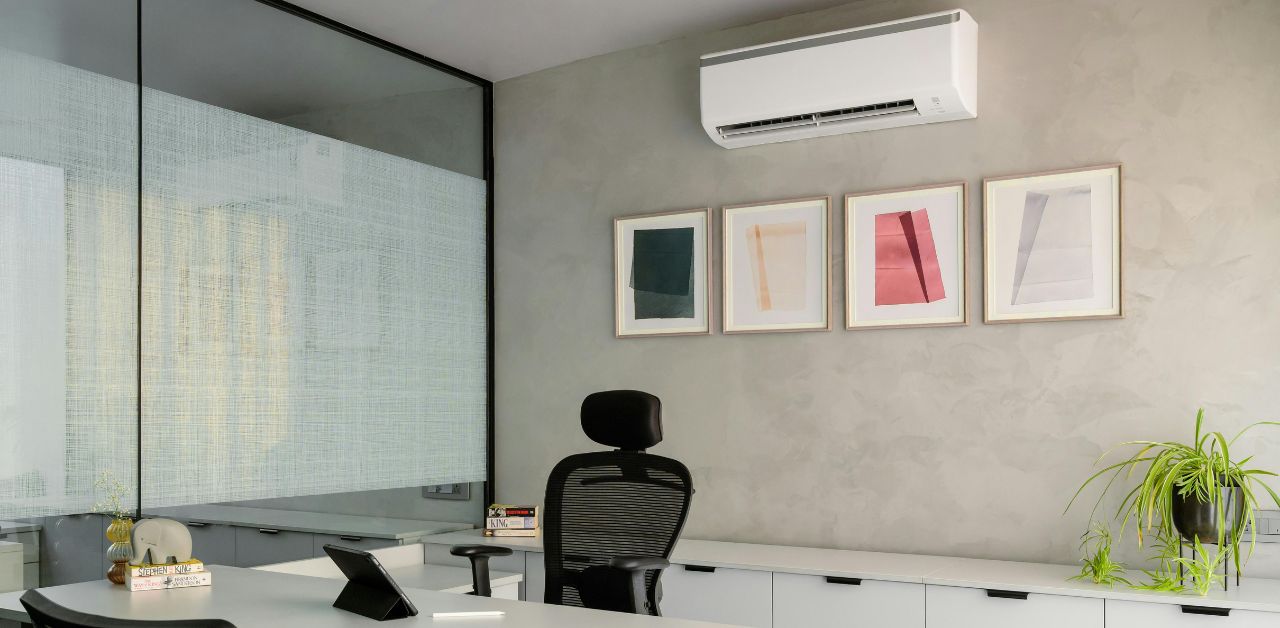 A small office with a desk and black computer chair. Behind it are 4 framed pictures and a split type air conditioner on top of the frames. This image depicts how Commercial Air Conditioning for Small Offices are installed.