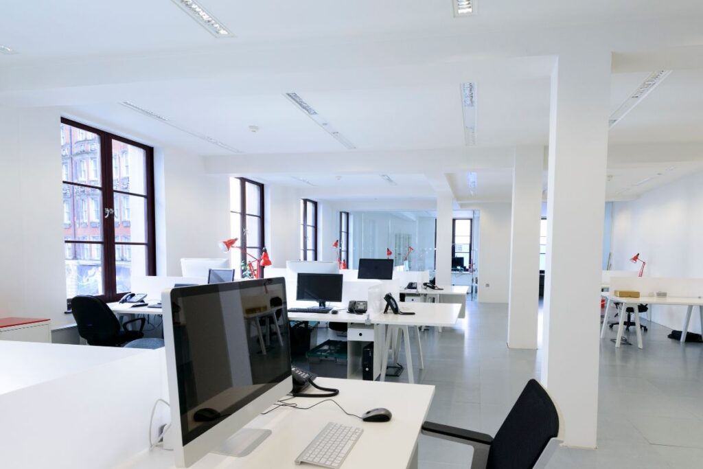 A small office space with 3 rows of desks with about 2 desktop sets each. The office is painted with wide windows to the left and foundational pillars on the right. This depicts how Commercial Air Conditioning for Small Offices are setup. 