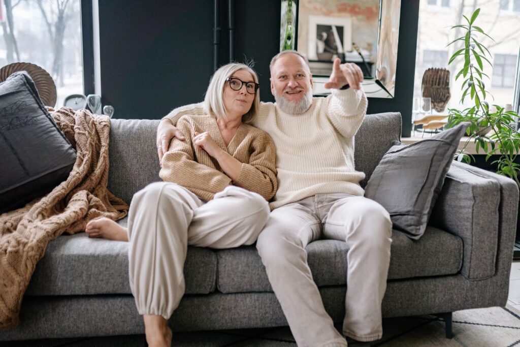 An elderly couple in jumpers and pants suits sitting in a gray couch. 